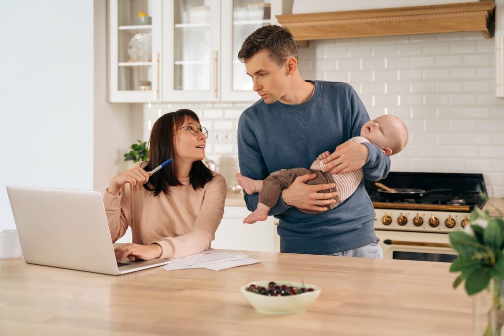 Young family with sleeping infant baby using laptop in the kitchen